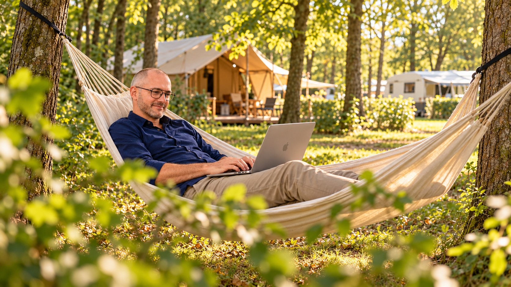 A man working on a laptop while relaxing in a hammock between trees at a wooded campsite